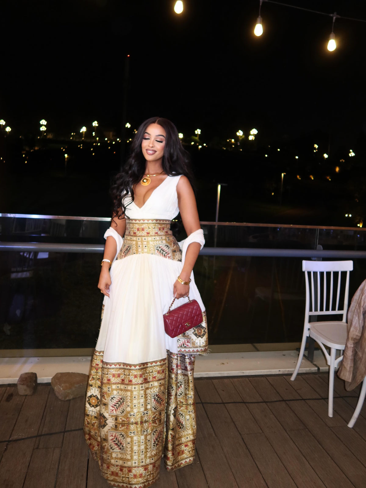 Woman in a white and gold Habesha Dress standing on a wooden deck at night.