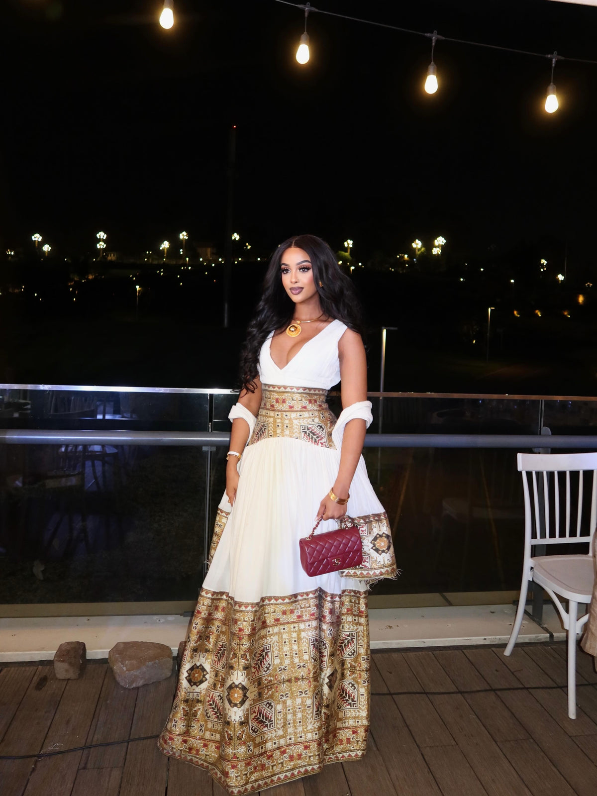 A woman stands on a deck at night wearing a white sleeveless Habesha Dress with a heavily embroidered gold and brown patterned waistband and hem. She is holding a small burgundy quilted handbag.