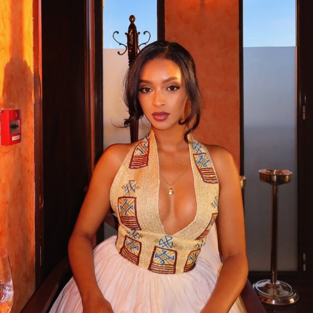 A woman wearing a halter-neck Ethiopian Clothing with a deep plunging neckline, decorated with geometric patterns in brown, blue, and gold on a beige base, paired with a white skirt. She is posed indoors against a warm-toned background.