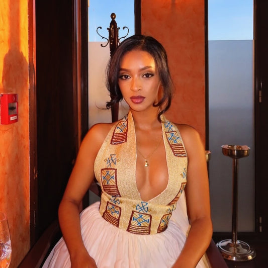 A woman wearing a halter-neck Ethiopian Clothing with a deep plunging neckline, decorated with geometric patterns in brown, blue, and gold on a beige base, paired with a white skirt. She is posed indoors against a warm-toned background.