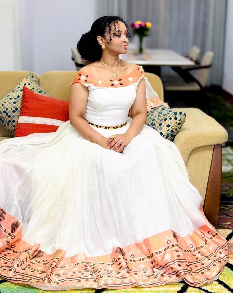 Woman in a white and orange traditional Habesha Kemis sitting on a couch.