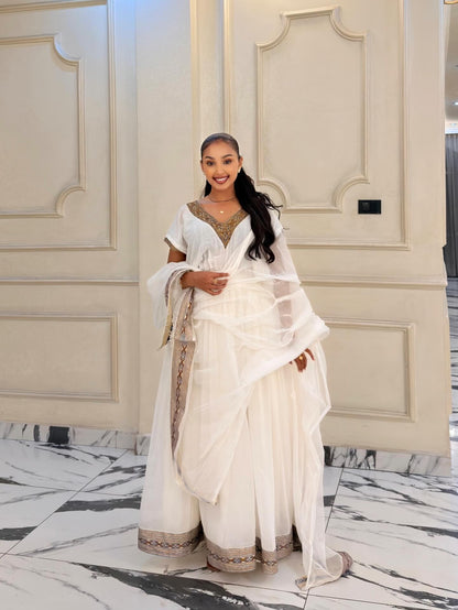 A woman wearing a white traditional Habesha Kemis with gold embroidery and a sheer white dupatta, standing in front of a cream-colored paneled wall and marble floor.
