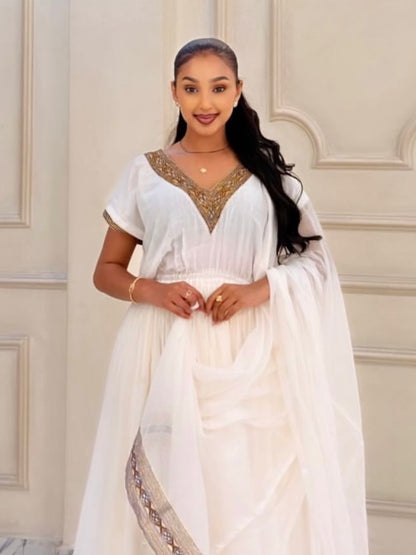 A woman wearing a white traditional Habesha Kemis with gold embroidered details on the neckline and a matching sheer white dupatta, standing against a cream-colored paneled wall.