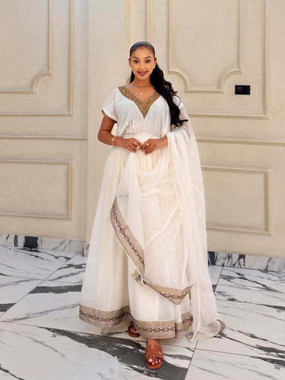 A woman modeling a white traditional Habesha Kemis with gold embroidery, standing in front of a beige paneled wall and marble floor.