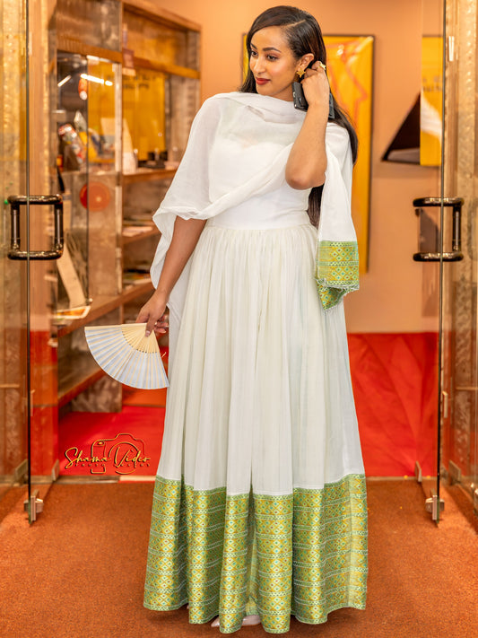 A woman models a traditional white maxi Ethiopian Dress with a green patterned hem and matching draped dupatta, holding a white folding fan in a warm-toned indoor setting.