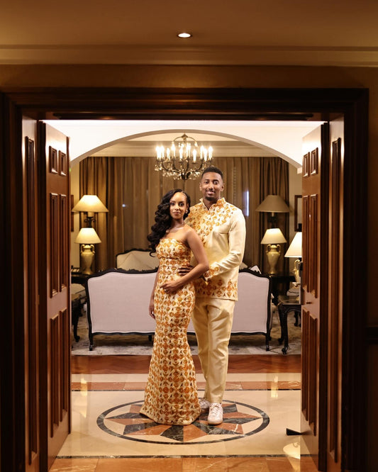 A couple posing in matching traditional African Ethiopian matching couple set  inside a luxurious hotel suite. The woman wears a strapless, floor-length beige dress with a brown geometric pattern, and the man wears a matching beige shirt and trousers with a brown geometric print.
