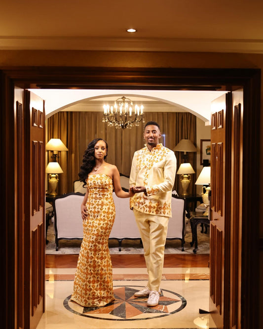 A couple posing in traditional Ethiopian matching couple set  inside a warmly lit hotel lobby. The woman wears a strapless, floor-length beige and gold patterned gown, and the man wears a matching beige and gold patterned shirt and trousers.