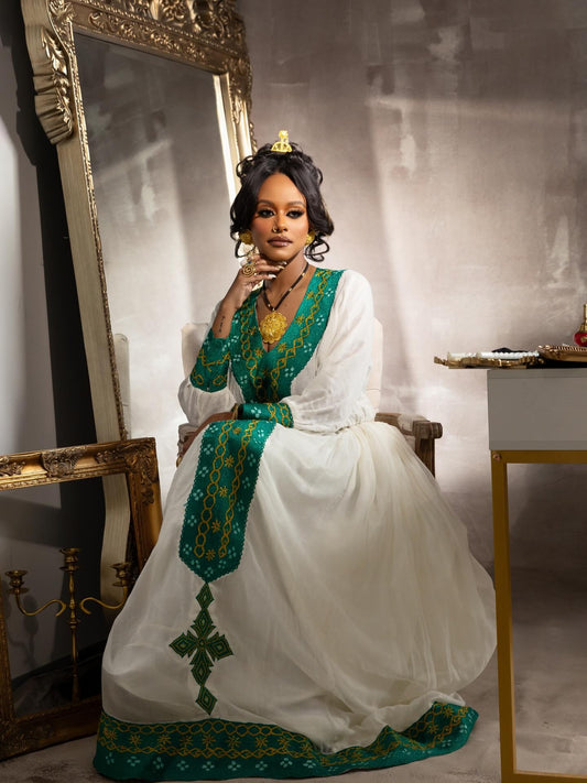 A woman seated in a vintage-style room wearing a traditional white Habesha Kemis with rich green and gold embroidery, a matching green sash, and a gold crown. She is positioned in front of a large ornate gold mirror.