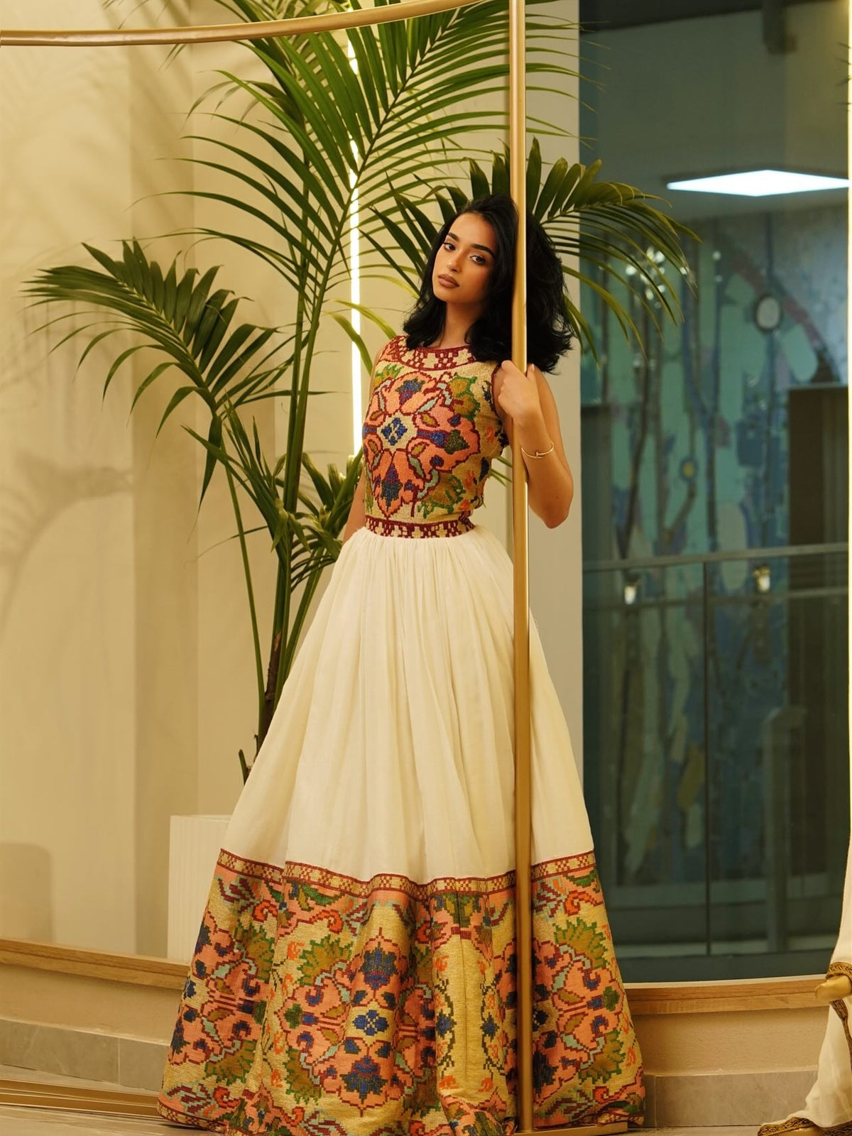 A woman modeling a traditional Indian Habesha Dress  with a cream skirt and a heavily embroidered multicolor bodice and hem, standing indoors next to a palm plant and a vertical wooden pole.