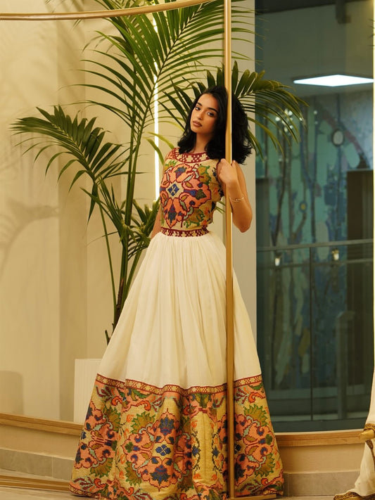 A woman modeling a traditional Indian Habesha Dress  with a cream skirt and a heavily embroidered multicolor bodice and hem, standing indoors next to a palm plant and a vertical wooden pole.