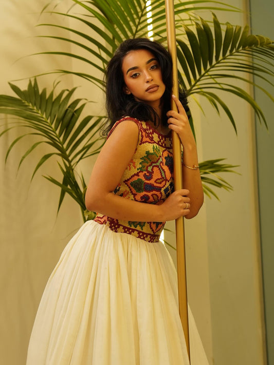 A woman posing in a sleeveless Habesha Dress  with a colorful, patterned embroidered bodice and a voluminous cream tulle skirt, holding a wooden pole in front of a warm beige wall with green palm leaves.