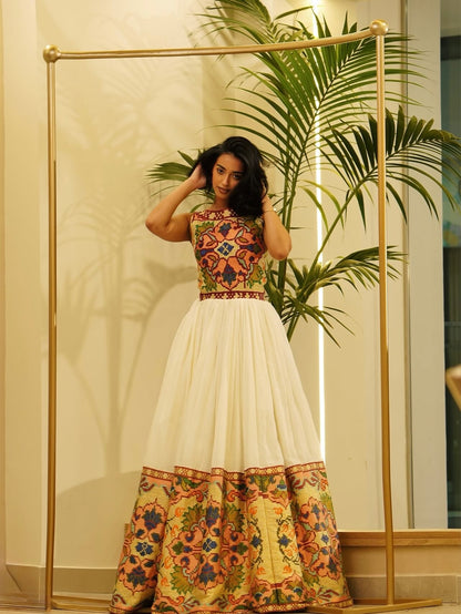 A woman modeling a sleeveless Habesha Dress  with a colorful geometric patterned bodice and a white flared skirt featuring a floral border print, standing in front of a gold clothing rack and a potted palm.