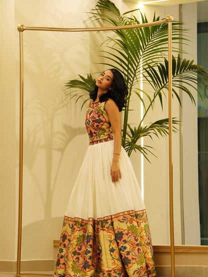 A woman models a sleeveless white Habesha Dress s with a colorful floral printed bodice and a matching floral printed skirt, standing in front of a gold clothing rack and a potted palm.