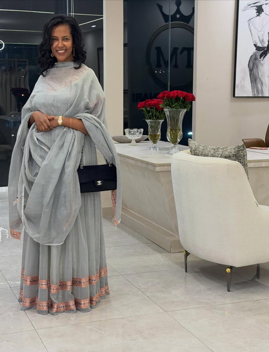 A woman wearing a light gray traditional Traditional Ethiopian Clothing with peach border detailing and a matching dupatta, standing in a modern boutique setting.
