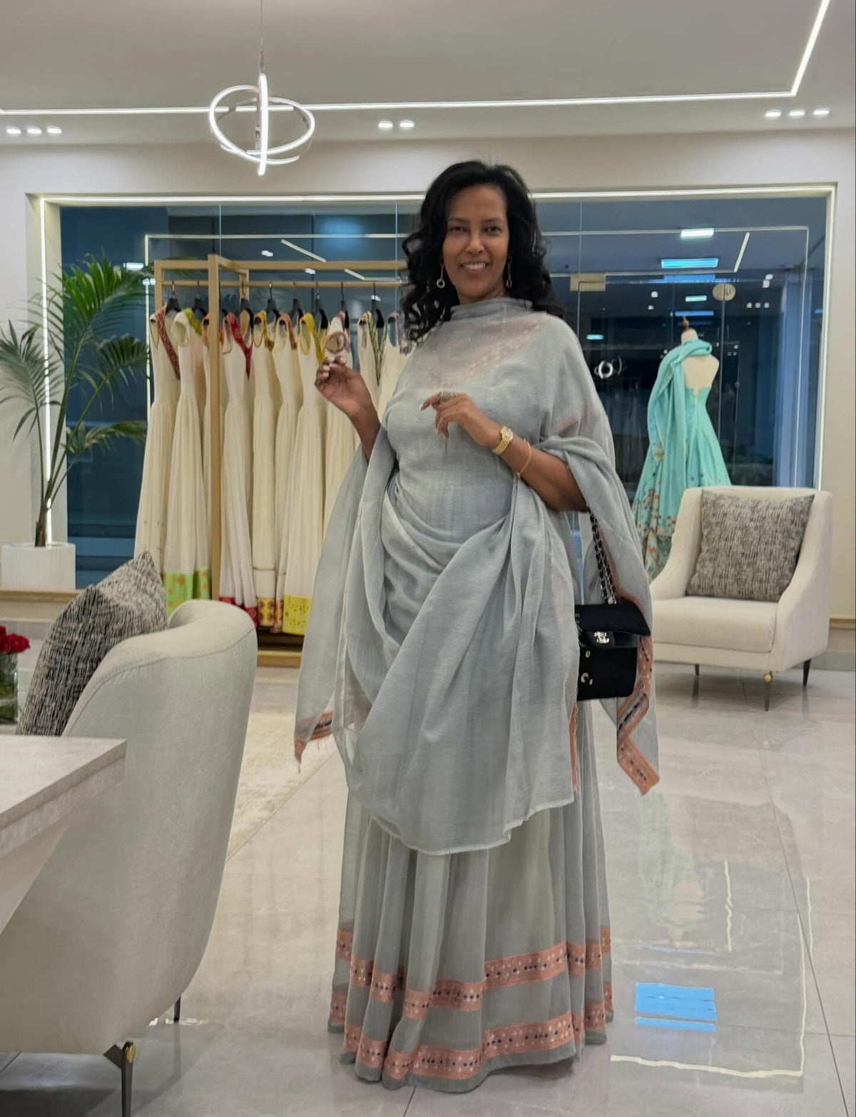 A woman posing in a modern boutique wearing a light gray traditional Traditional Ethiopian Clothing with a matching dupatta and a peach-colored embroidered border. The background shows other garments on racks and a mannequin wearing a light blue dress.