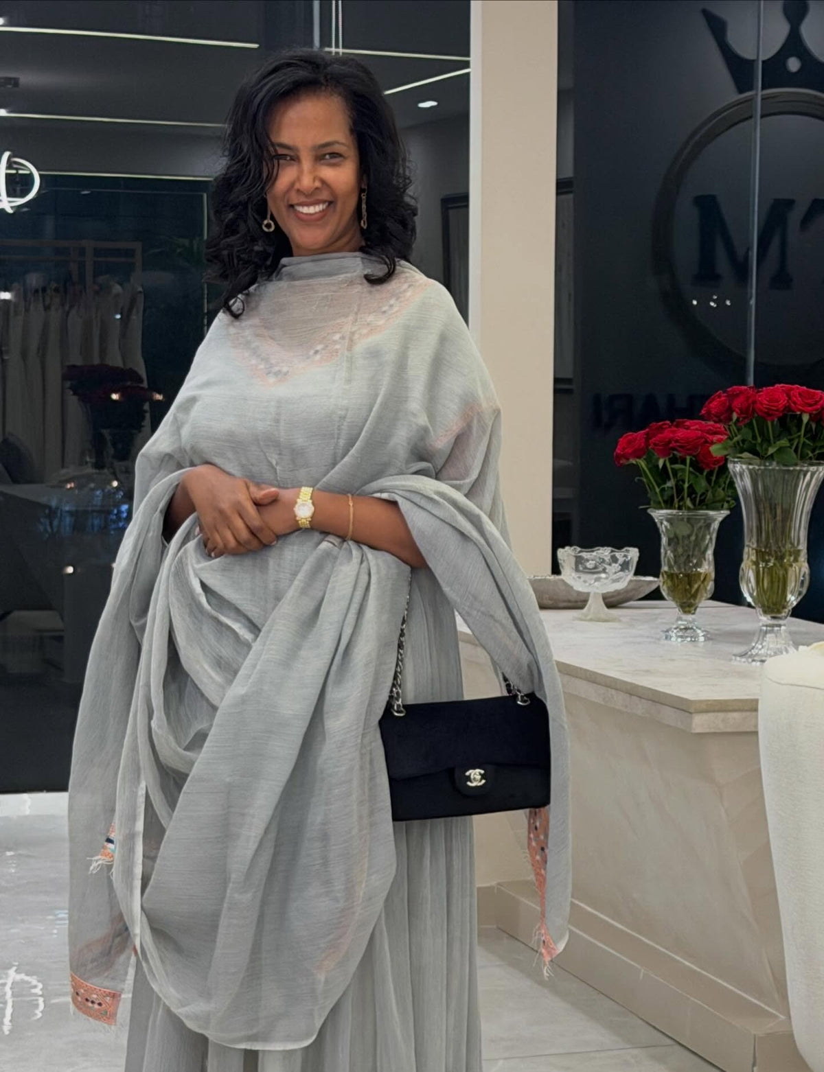 A woman smiling while wearing a light gray draped Traditional Ethiopian Clothing with a matching sheer shawl and holding a black Chanel shoulder bag in a modern indoor setting.