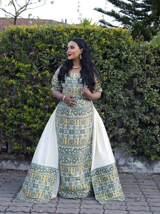 A woman wearing a traditional long Habesha Wedding Dress with green and beige geometric patterns and a white draped overlay, standing outdoors in front of a green hedge.