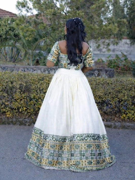 A woman viewed from behind wearing a traditional Habesha Wedding Dress with a white skirt and a green and yellow embroidered blouse, standing outdoors on a paved path.
