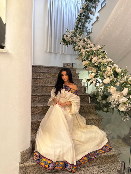 Woman in a white Habesha kemis with colorful embroidery sitting on a staircase.