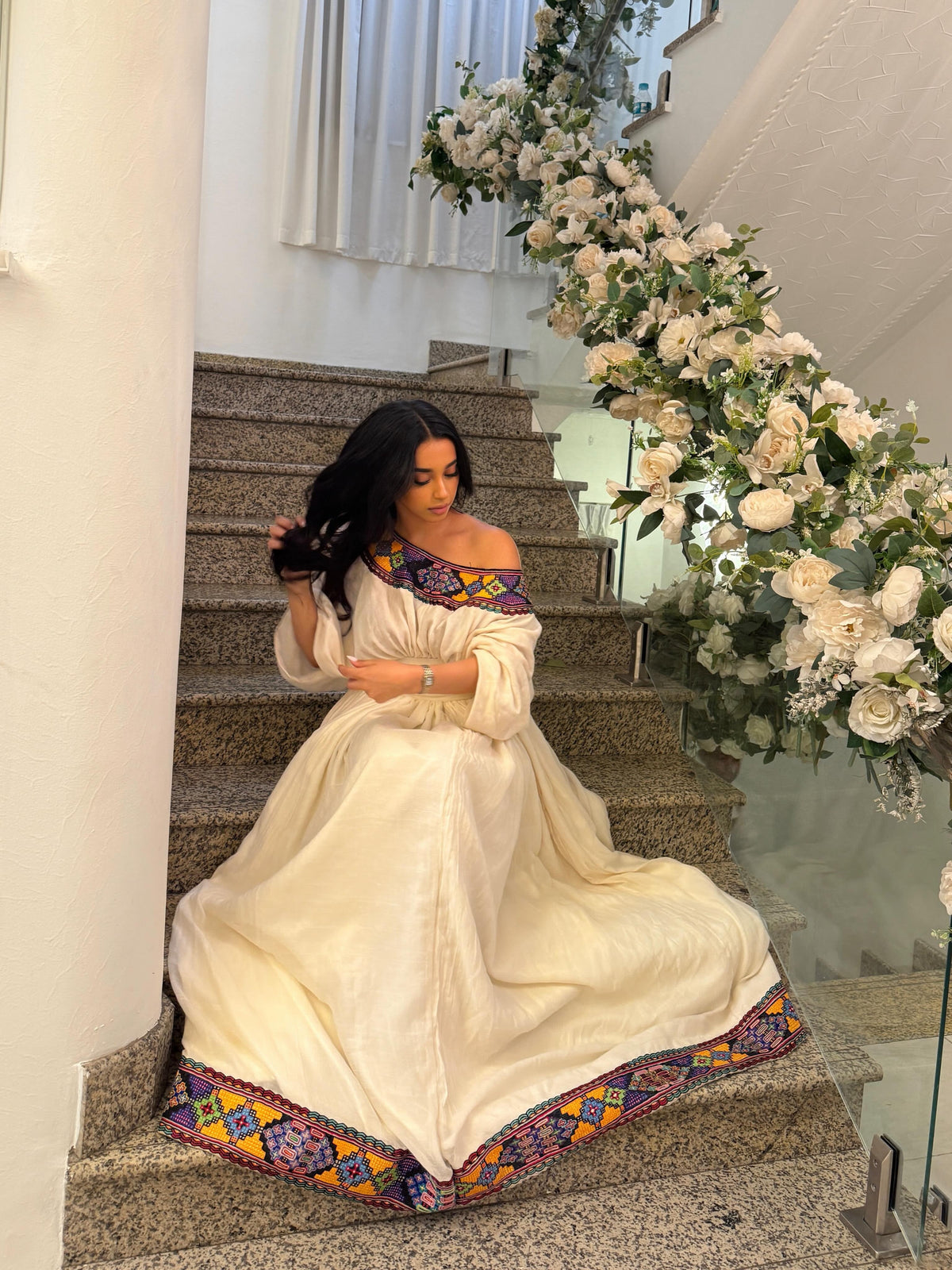 Woman in a white traditional Habesha kemis with colorful patterns sitting on stairs next to floral decorations.