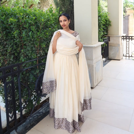 Woman in a white traditional Habesha dress with black patterns standing outdoors.