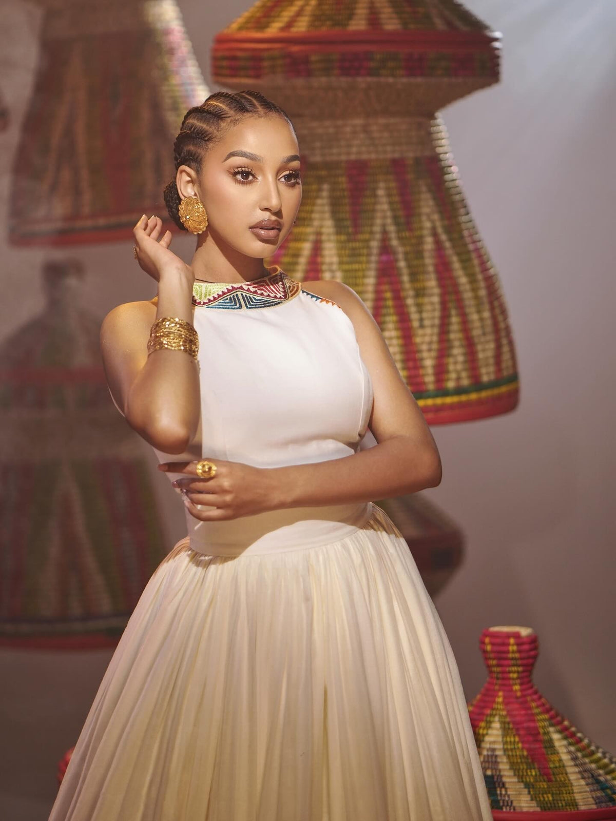 A woman models a sleeveless white Habesha Dress with a pleated skirt, styled with gold jewelry and a colorful beaded collar, set against a background of woven baskets.