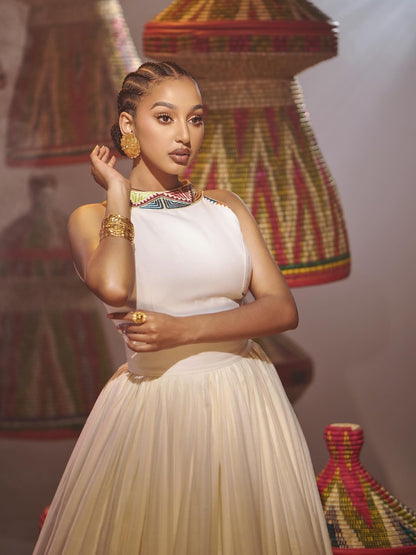 A woman models a sleeveless white Habesha Dress with a pleated skirt, styled with gold jewelry and a colorful beaded collar, set against a background of woven baskets.