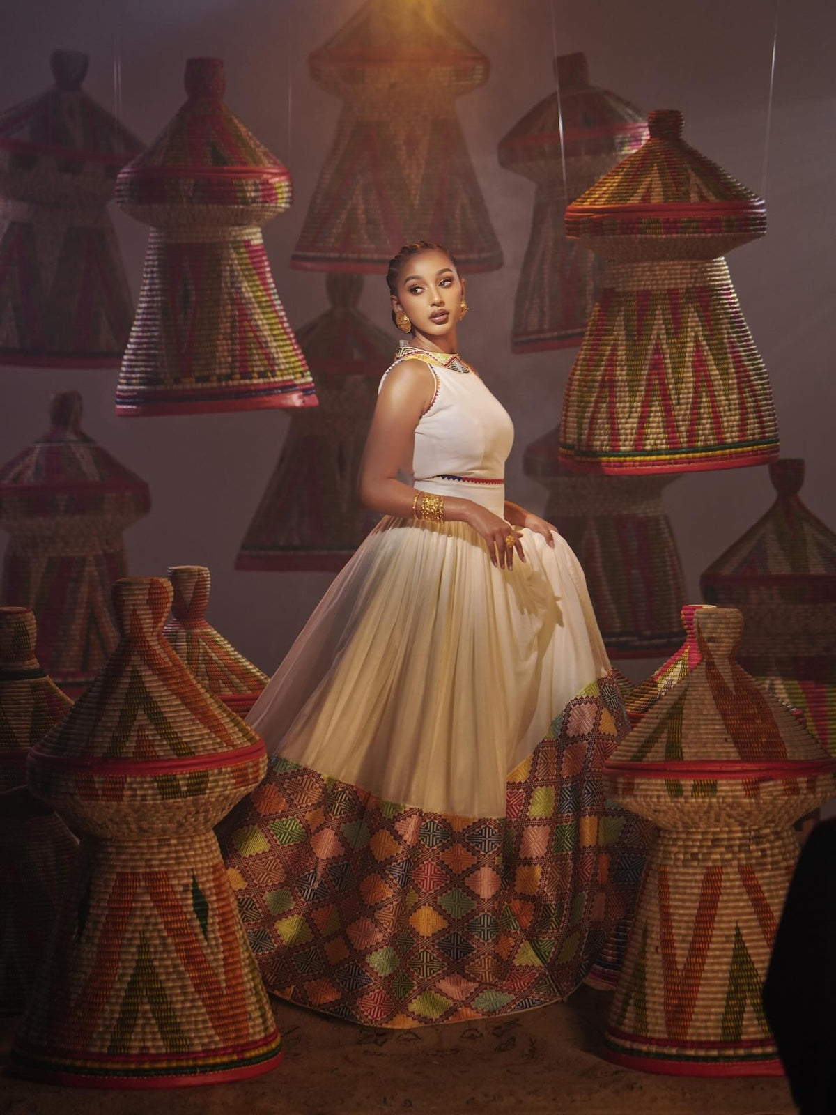A woman stands in a studio wearing a sleeveless Habesha Dress with a patterned multicolor hem, surrounded by large woven cone-shaped baskets in matching earthy tones.