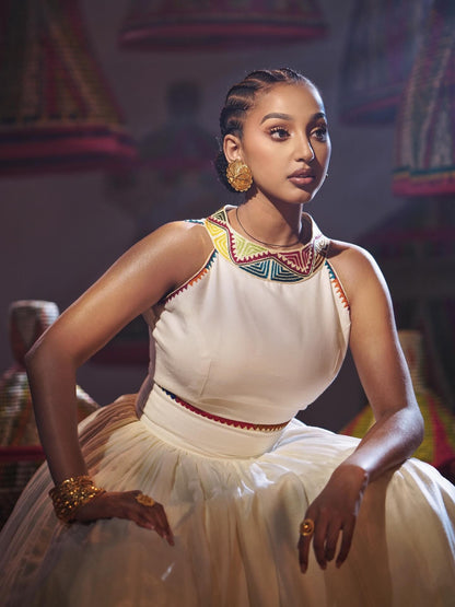 A woman wearing a sleeveless white Habesha Dress with colorful geometric embroidery around the neckline and waist, posed in a dimly lit, ornate interior.