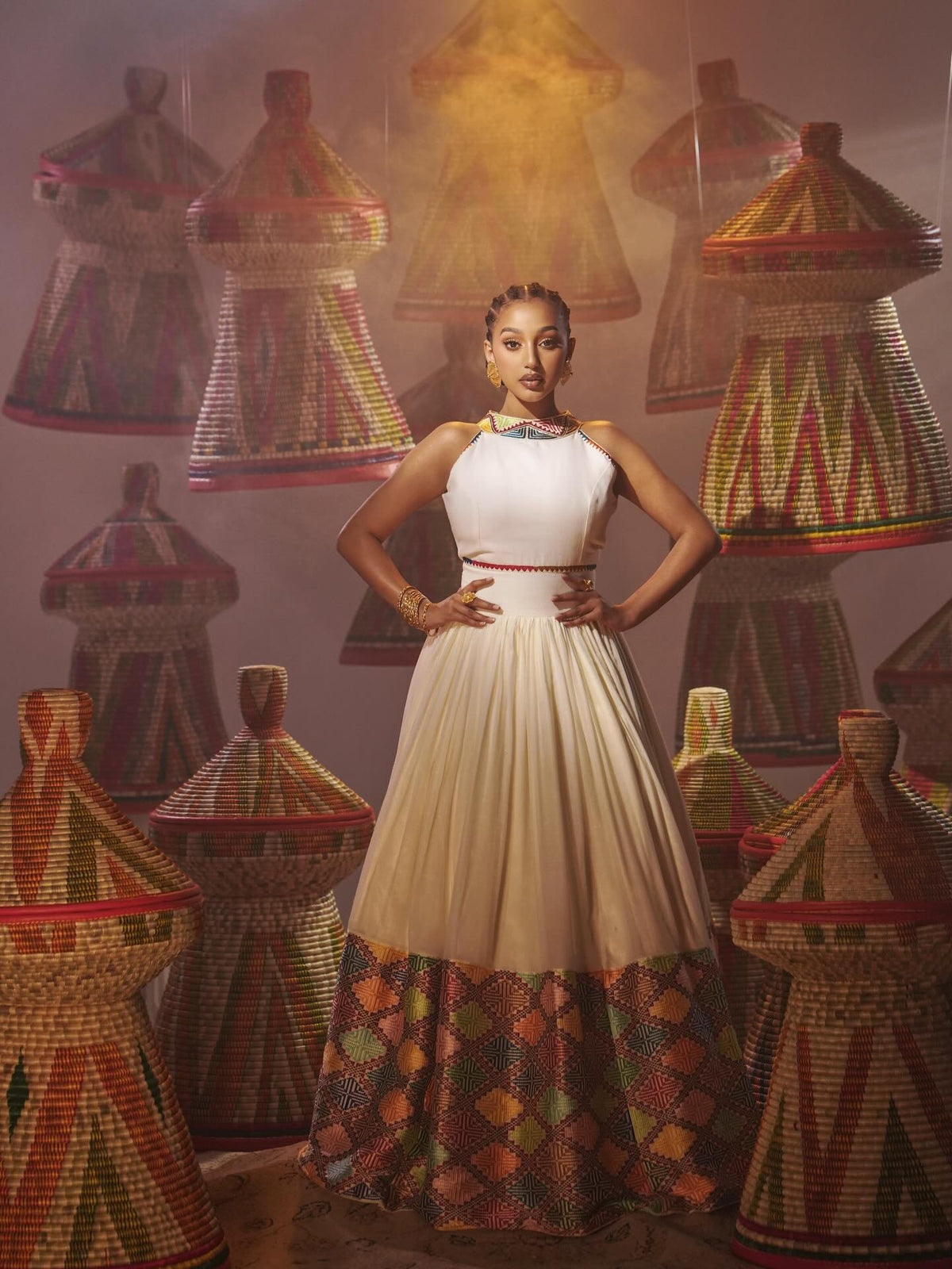 A woman stands in a studio wearing a sleeveless cream Habesha Dress with a patterned multicolor hem, surrounded by hanging woven cone-shaped garments in warm golden and orange tones.