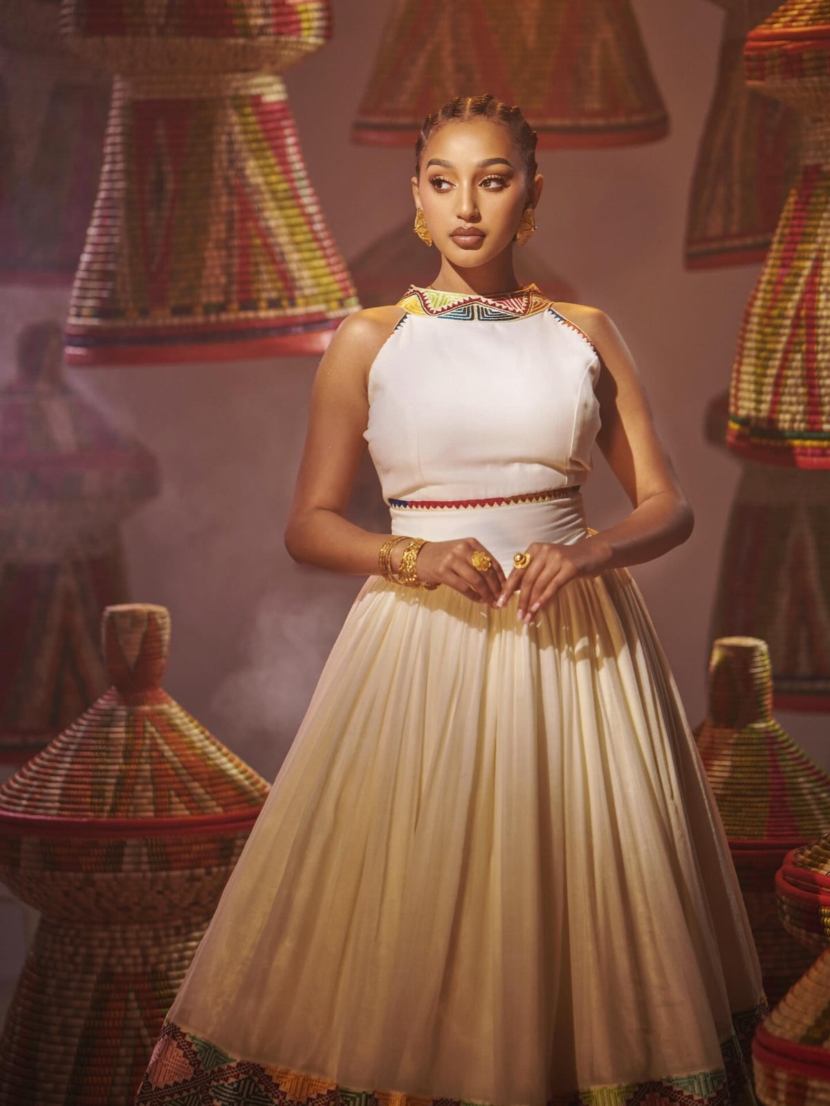 A woman stands in a studio wearing a sleeveless Habesha Dress with gold jewelry, surrounded by woven decorative baskets.