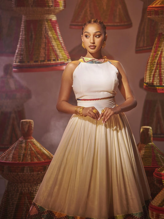 A woman stands in a studio wearing a sleeveless Habesha Dress with gold jewelry, surrounded by woven decorative baskets.