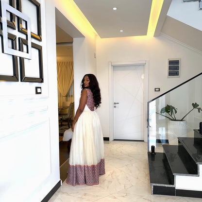 A woman wearing a sleeveless white Ethiopian Dress with a purple patterned hem and shoulder detailing, standing in a modern white hallway with marble floors and a staircase.