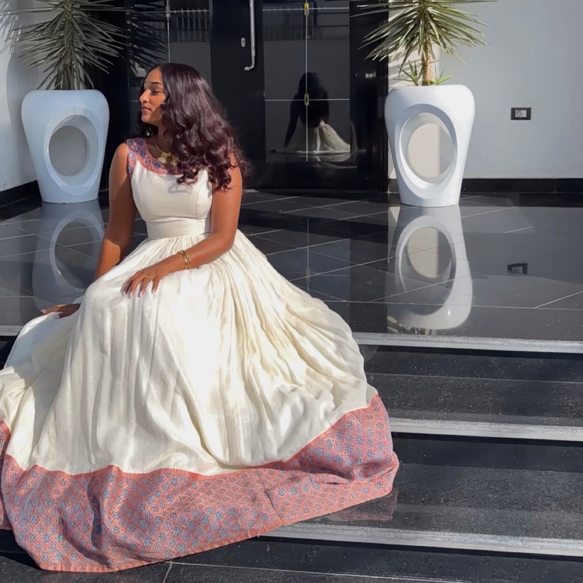 A woman wearing a voluminous cream-Ethiopian Dress with a patterned pink and blue border sits on black steps in a modern lobby.