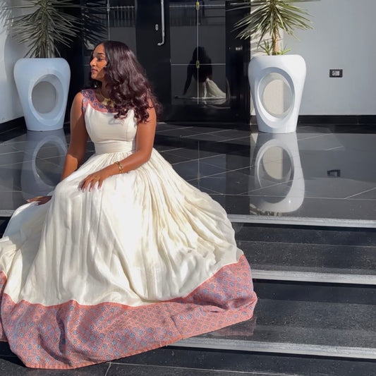 A woman wearing a voluminous cream-Ethiopian Dress with a patterned pink and blue border sits on black steps in a modern lobby.