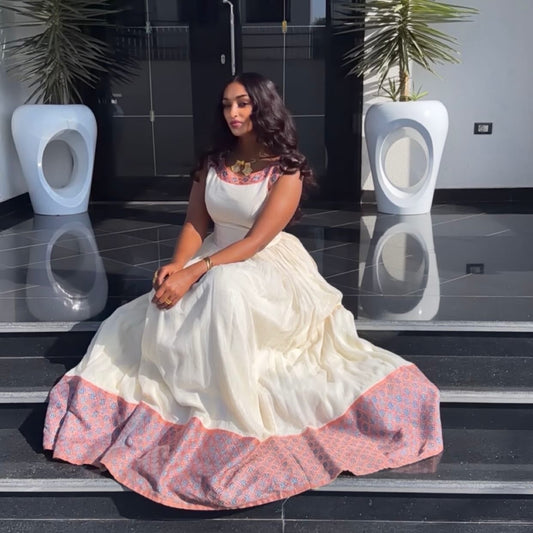 A woman wearing a voluminous white or cream Ethiopian Dress with pink patterned trim sits on black steps in front of a modern building entrance. The dress has a solid cream body with a wide pink and blue patterned border at the hem and a matching pink patterned neckline.