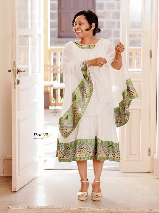 A woman modeling a white traditional Habesha Dress with green and beige patterned borders and dupatta, standing in a doorway.