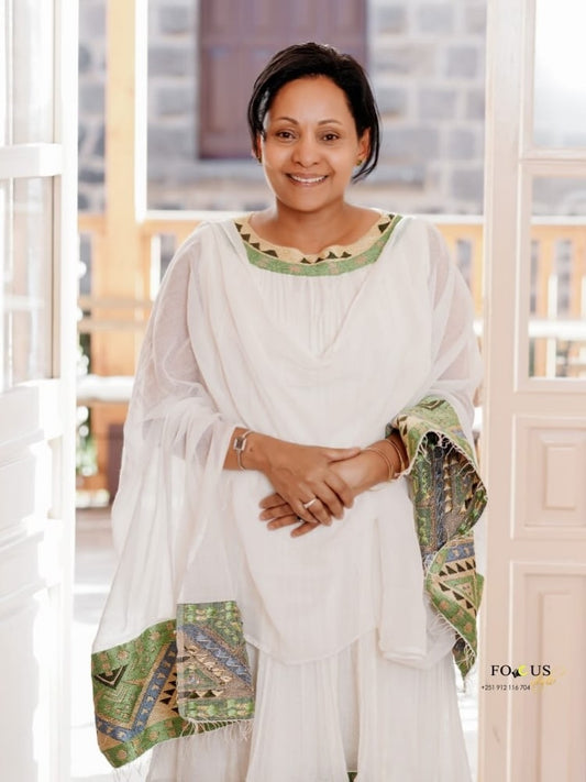 A woman wearing a white traditional Habesha Dress with green and gold patterned trim, standing in front of a bright doorway.