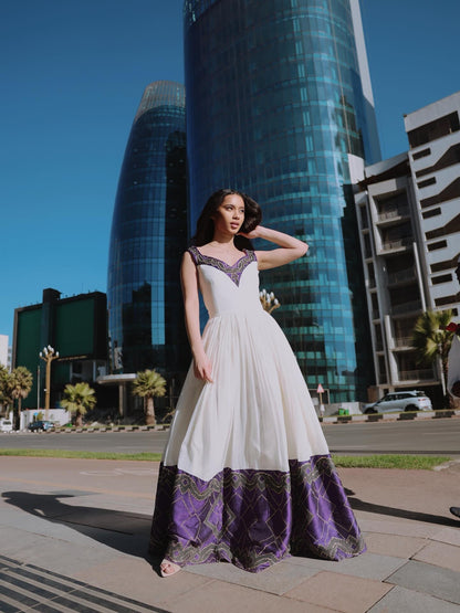 A woman wearing a white and purple Habesha Dress stands on a city sidewalk with modern glass skyscrapers in the background under a clear blue sky.