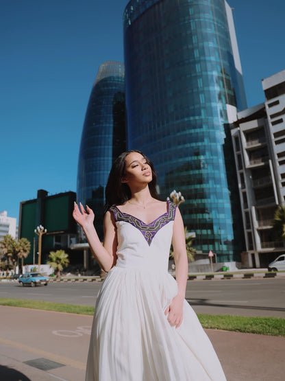 A woman wearing a white sleeveless A-line Habesha Dress with a purple and gold embroidered V-neckline stands outdoors in front of modern glass skyscrapers under a clear blue sky.