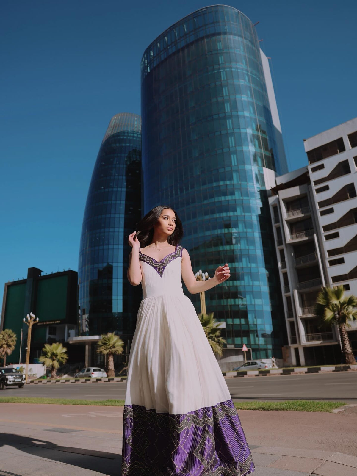 A woman wearing a white and purple traditional Habesha Dress stands outdoors in front of modern glass skyscrapers under a clear blue sky.
