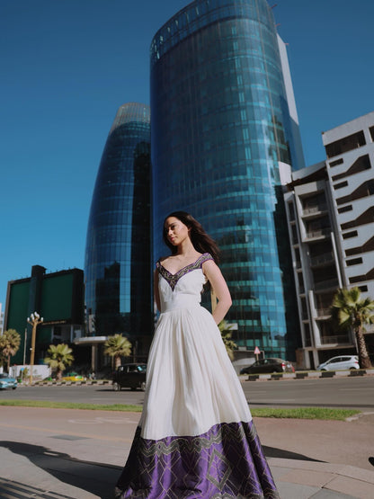 A woman wearing a white A-line Habesha Dress with purple and black patterned detailing at the neckline and hem, standing outdoors in front of modern glass skyscrapers under a clear blue sky.