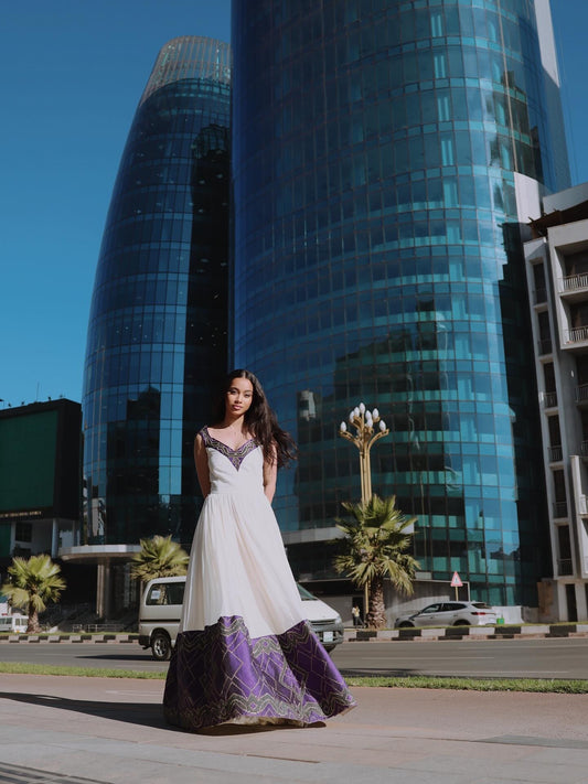 A woman wearing a white sleeveless Habesha Dress with a purple patterned skirt stands outdoors in front of two tall blue glass skyscrapers and palm trees.