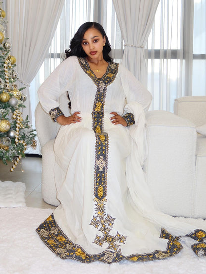 A woman wearing a white traditional Ethiopian Traditional Dress with ornate gold and dark blue embroidery, seated on a white sofa in a bright room with curtains, a Christmas tree, and a rug.