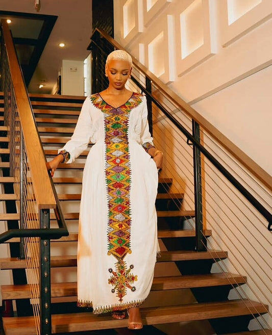 Woman in a long, white Habesha Kemis  with colorful embroidery standing on a staircase.