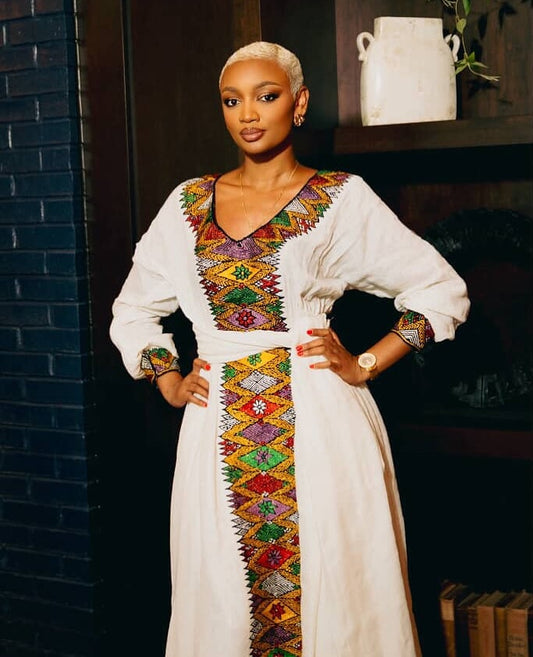 A woman modeling a white long-sleeve traditional Habesha Kemis  with vibrant multicolor geometric embroidery around the neckline, waist, and front opening, posed against a dark indoor background with a brick wall and wooden shelf.