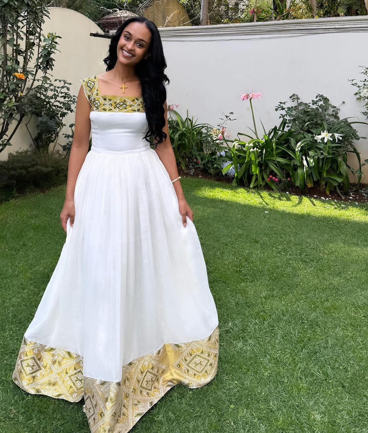 A woman stands on a green lawn wearing a white Habesha Dress  with gold and green patterned trim at the neckline and hem. The background shows a white wall, garden plants, and grass.