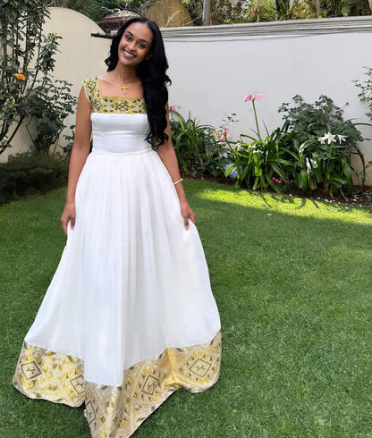 A woman stands on a green lawn wearing a white Habesha Dress  with gold and green patterned trim at the neckline and hem. The background shows a white wall, garden plants, and grass.