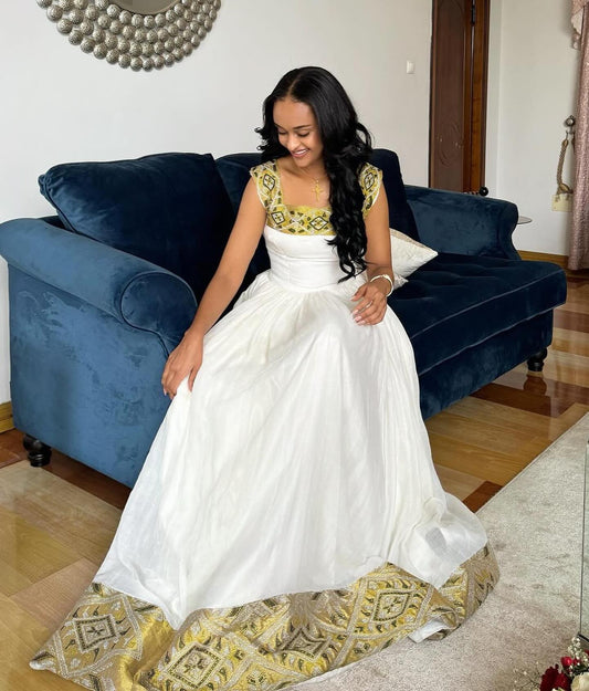 A woman wearing a white A-line Ethiopian Dresswith gold floral embroidery on the bodice, sitting on a dark blue velvet sofa in a living room.