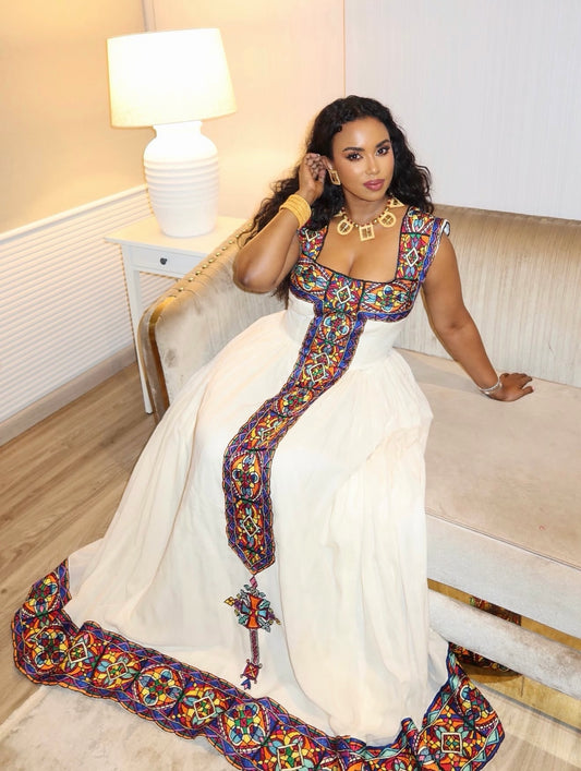 Woman wearing a white sleeveless Habesha kemis featuring colorful geometric Tilf embroidery along the square neckline, vertical front panel, and hem.